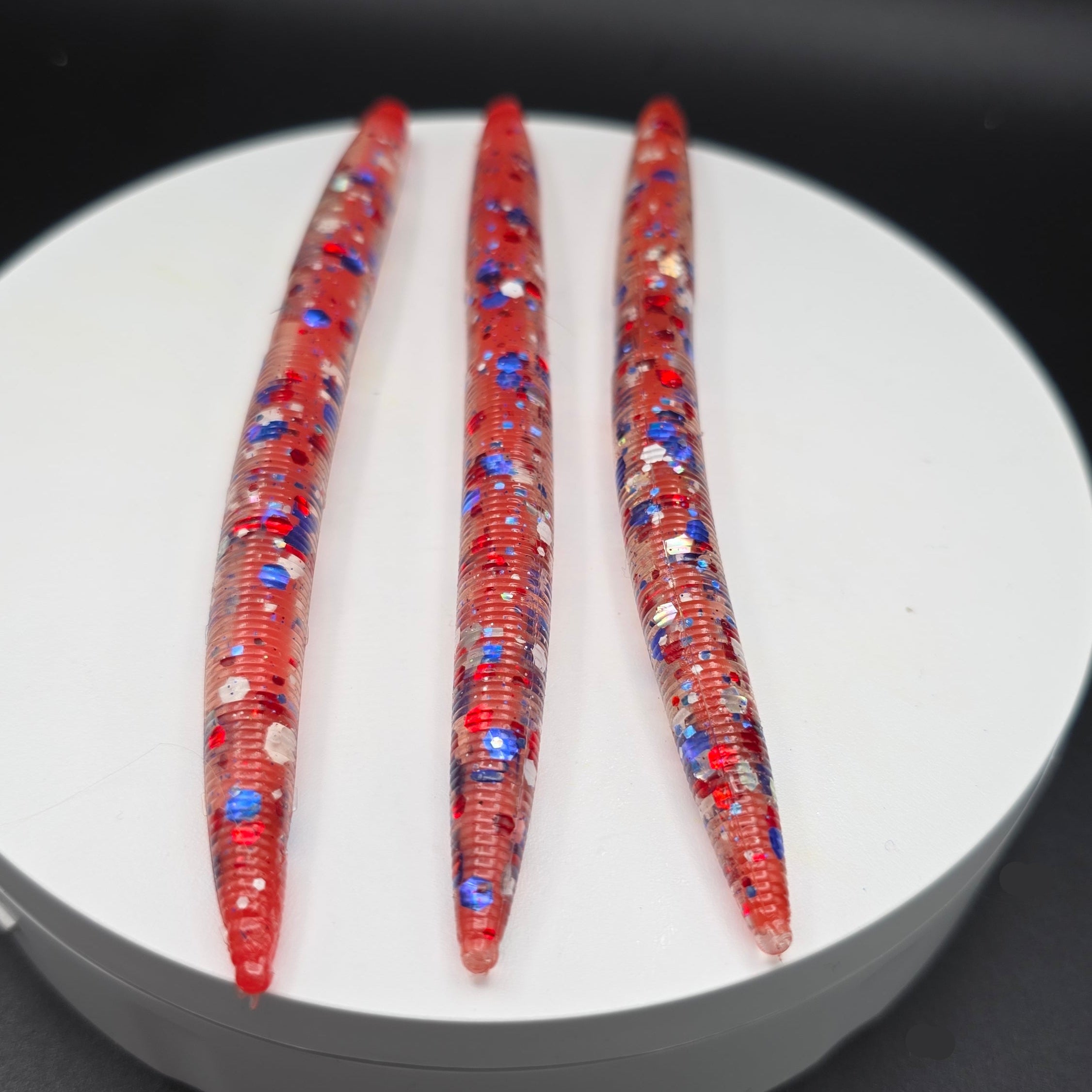 Three Clear with UV Red Core fishing lures with red, white, and blue glitter  on a white circular platform with a dark background